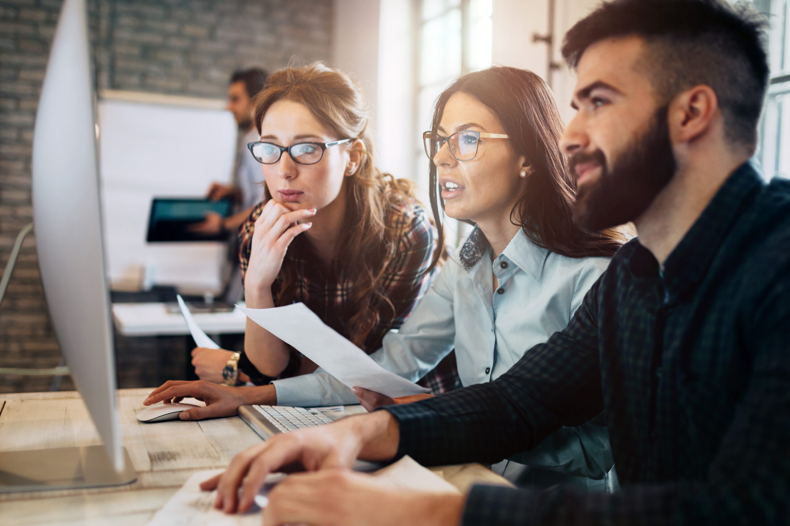 Three coworkers gathered around a desktop computer, reviewing documents in a bright office.