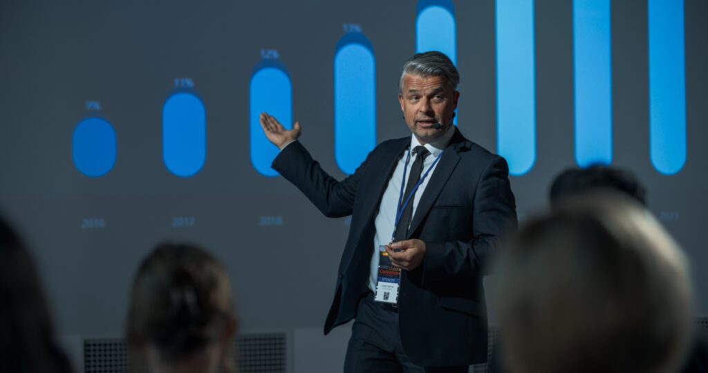 Male presenter in a dark suit giving a talk on stage, gesturing with his left hand, with a blue bar-chart backdrop behind him.